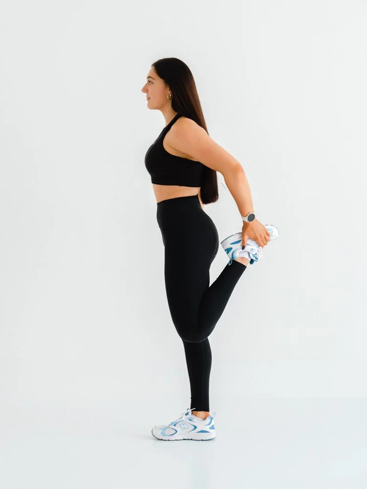 A woman in black athletic clothing doing a quadriceps stretch with her right leg bent behind her, holding her ankle with her right hand, in a minimally decorated white space.