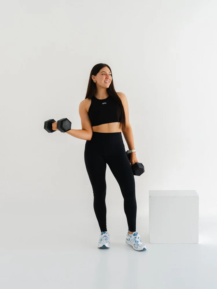 A woman in sportswear holding dumbbells, with a white background and a white cube in front of her.