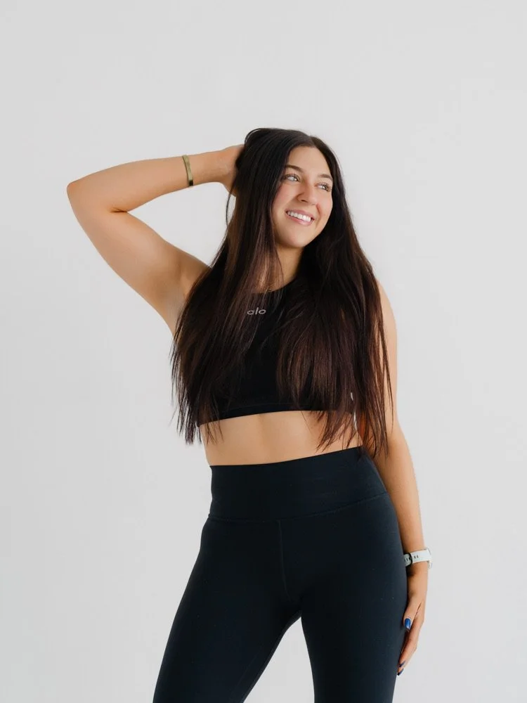 A young woman with long dark hair, wearing a black sports bra and black leggings, smiling and posing with one hand behind her head against a plain white background.