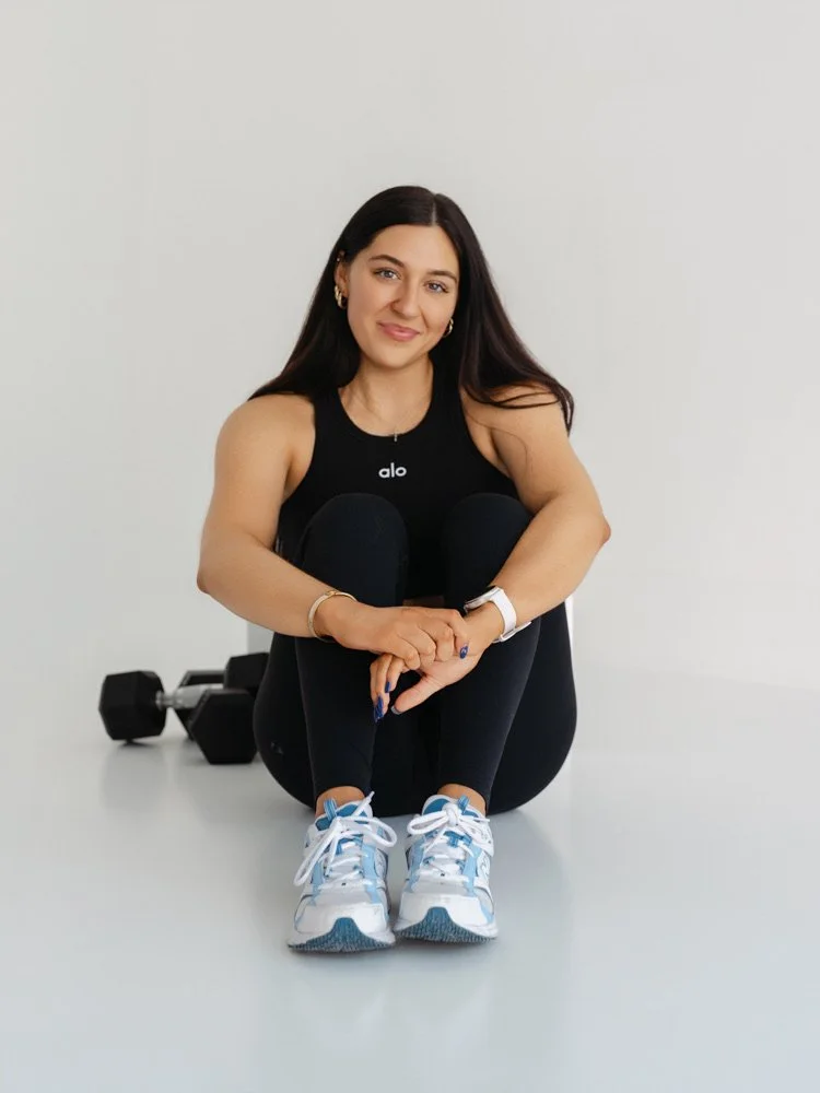 A woman sitting on the floor with her knees drawn up, wearing a black workout top, black leggings, and white sneakers. She has long dark hair and is smiling. There are dumbbells in the background.