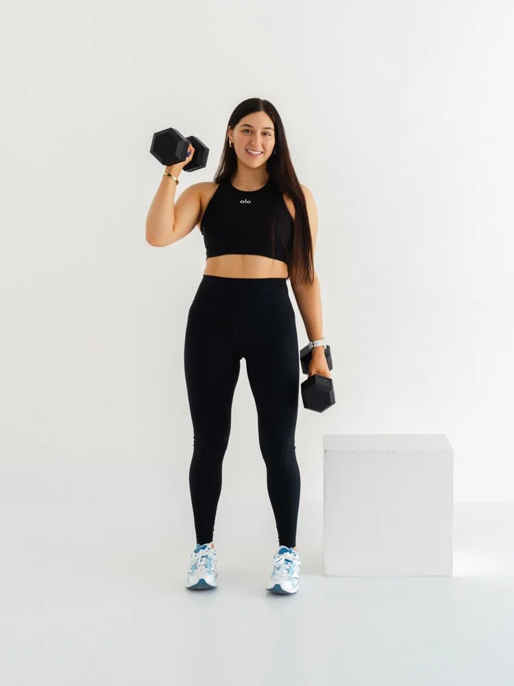 Fit woman in black athletic wear posing with dumbbells in a minimalist white studio.