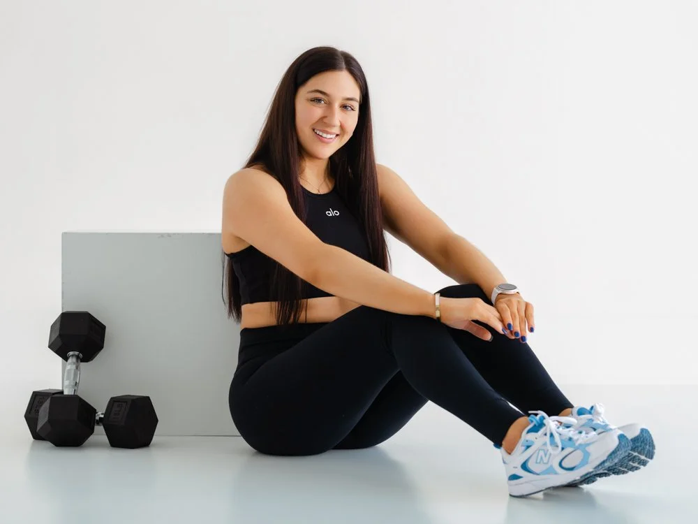 A woman in workout clothes sitting on the floor, smiling, with dumbbells and a fitness mat nearby.