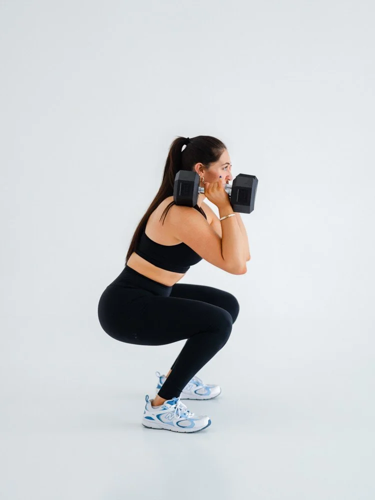 Woman in black workout clothes squatting while lifting a dumbbell with each hand in front of a plain white background.