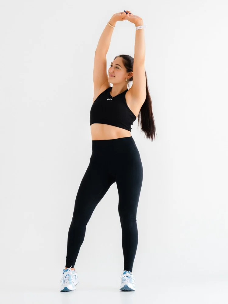 Woman in black workout outfit stretching with arms overhead in a fitness setting.