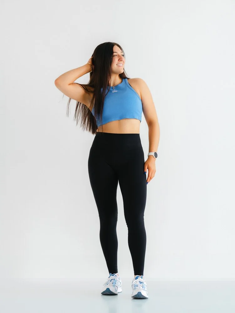 A young woman in athletic wear smiling with one hand behind her head standing against a plain white background.