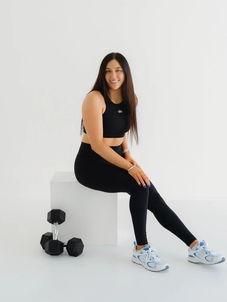 A woman with long dark hair sitting on a white block in a gym setting, smiling, wearing black athletic wear, sneakers, and fitness tracker, with dumbbells on the floor beside her.
