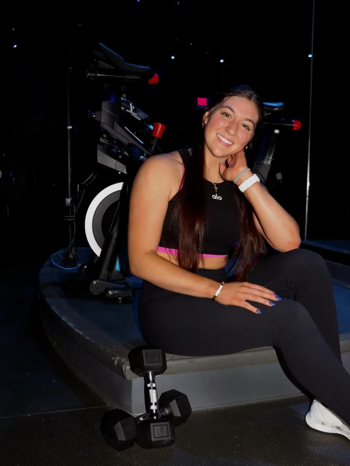 A young woman with long dark hair, wearing black workout clothes and a white wristband, sitting on a fitness machine in a dark gym, smiling at the camera.