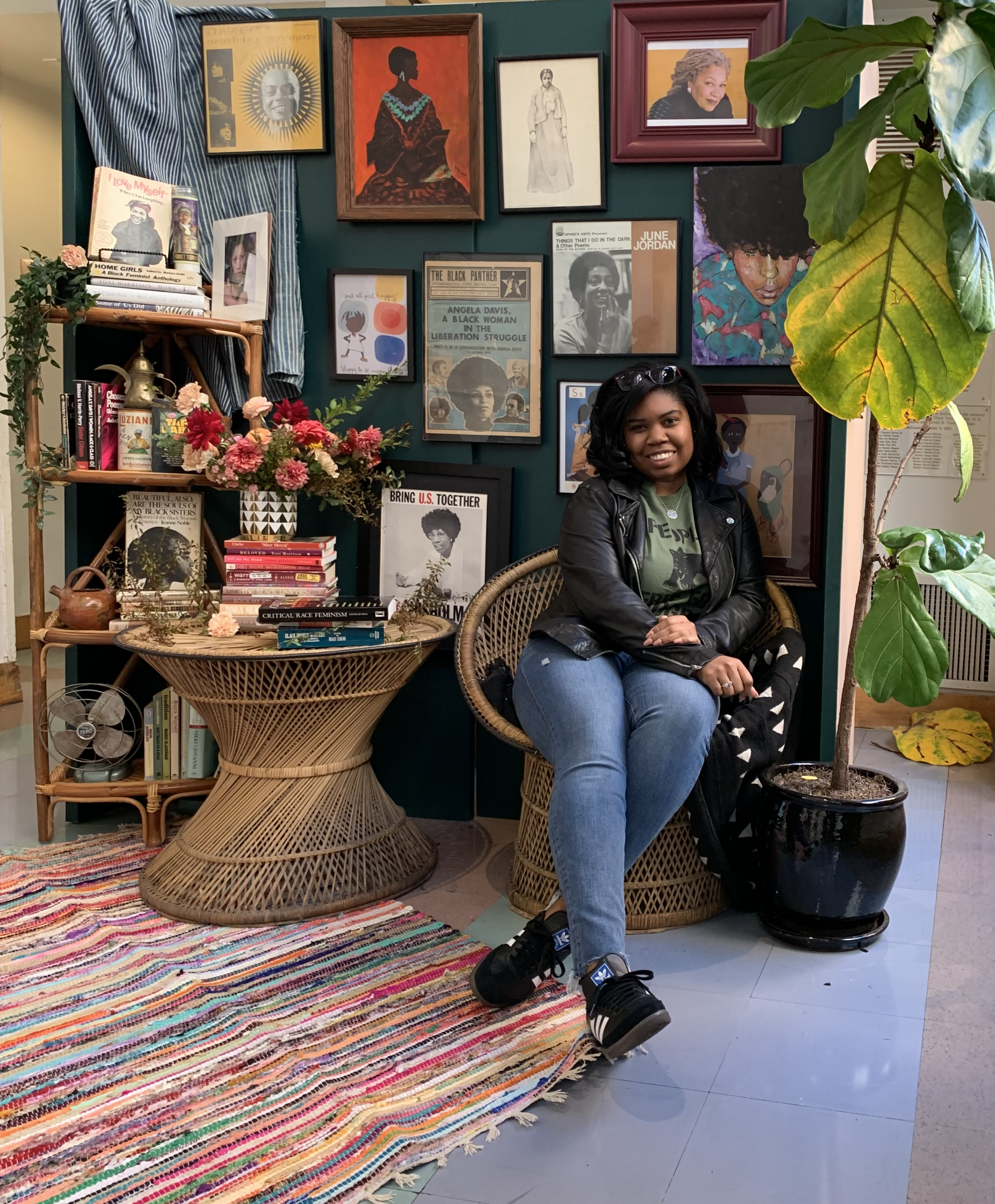 Young woman sitting on wicker chair in a colorful, art-filled room with framed pictures and posters of Black women and civil rights figures, a large potted fig tree, a multicolored rug, and a rattan table with books and flowers.