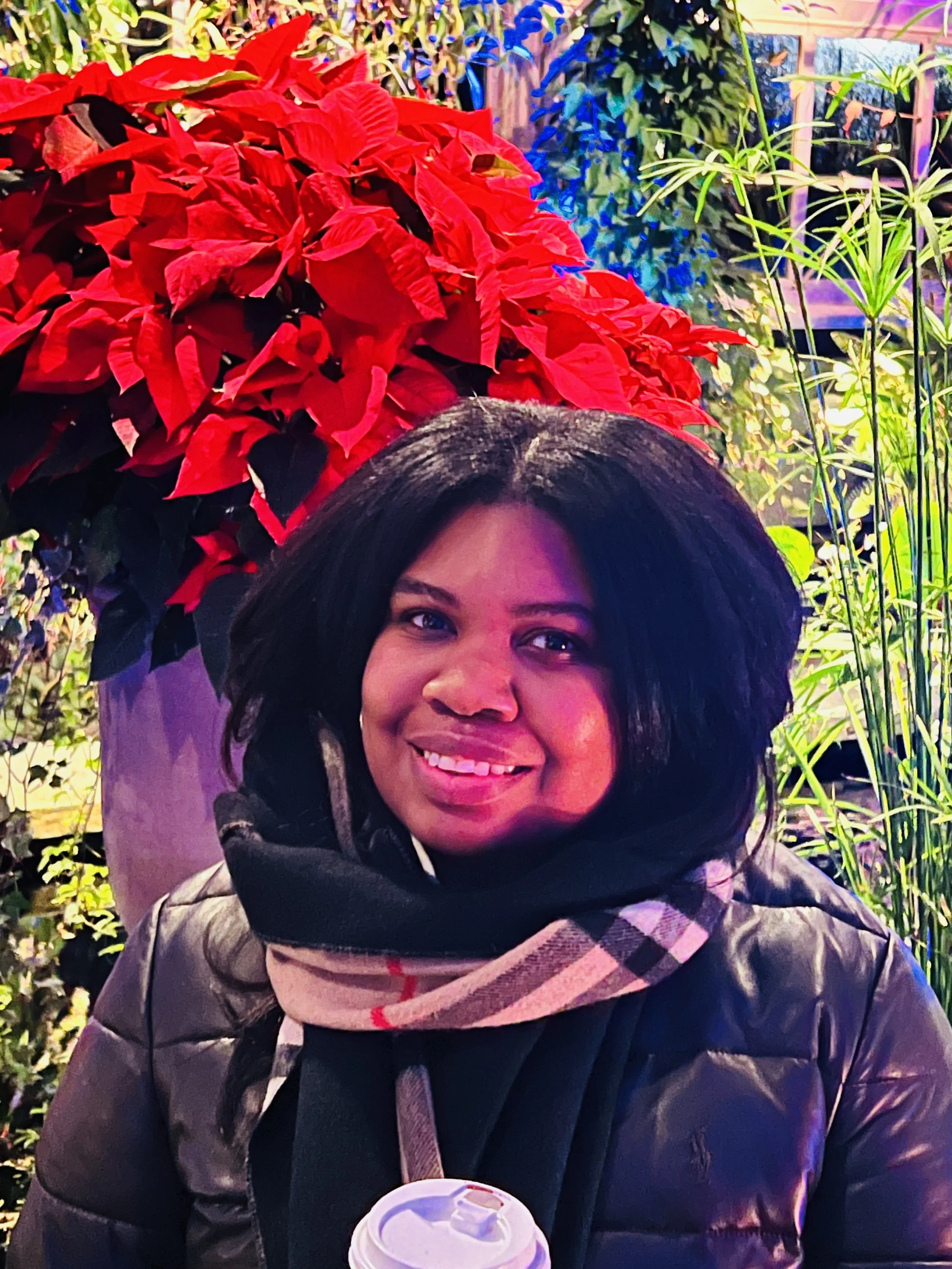 A smiling woman with dark hair standing in front of red poinsettia flowers and green plants, holding a cup with a white lid.