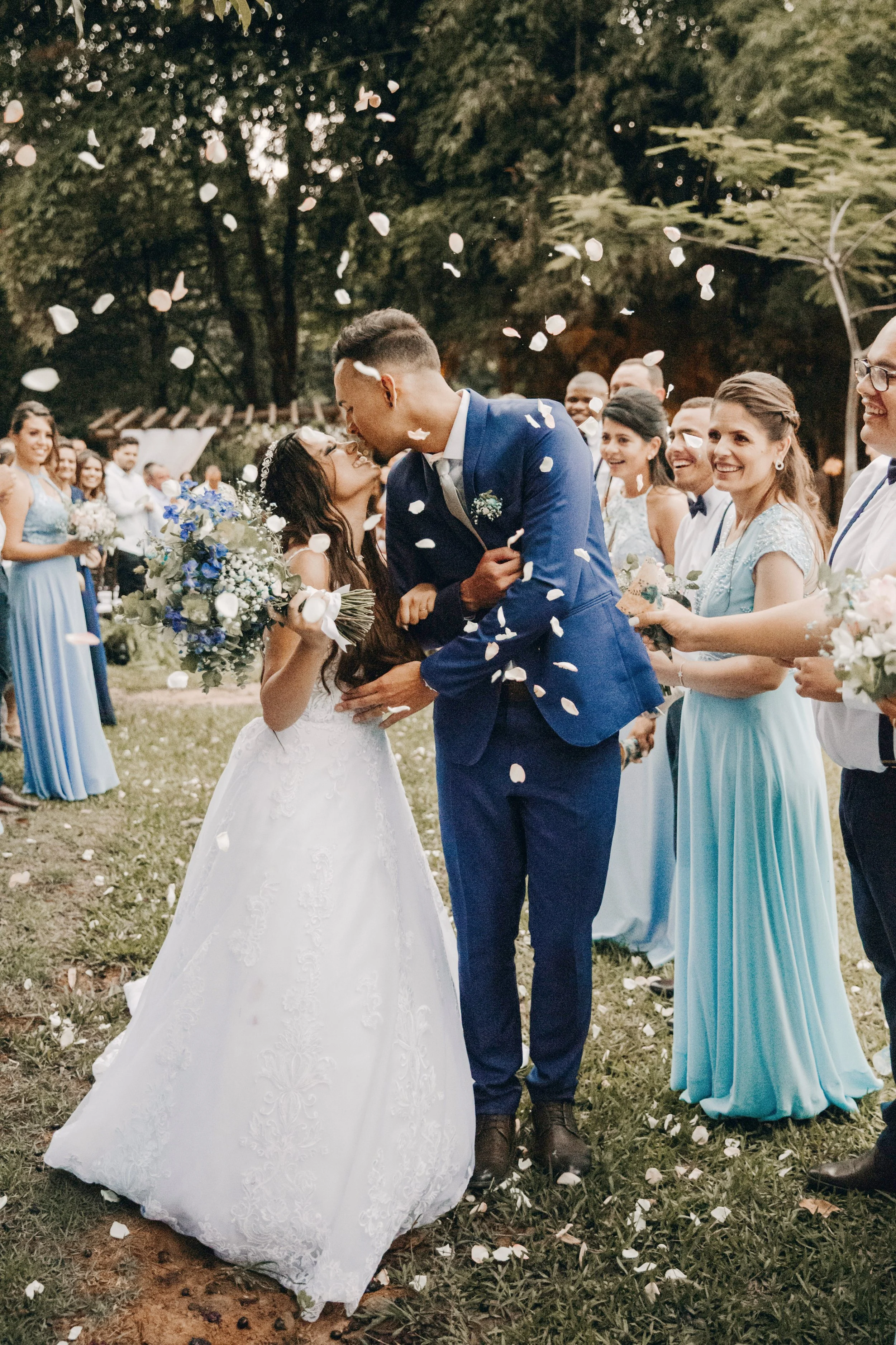 A wedding scene with a bride and groom surrounded by friends and family outdoors. Petals are falling as they kiss, and the guests are smiling and holding bouquets, dressed in formal attire.