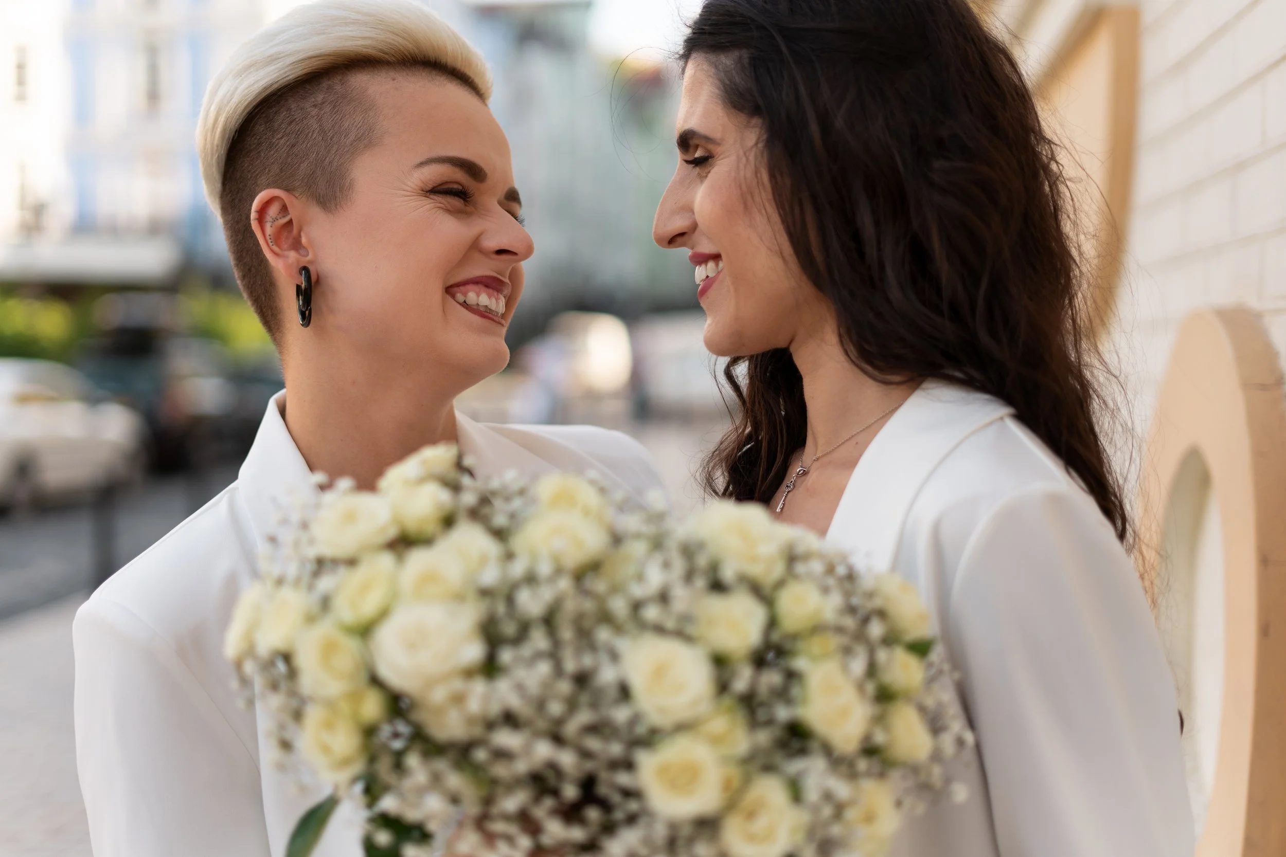 Two women smiling and facing each other outdoors, one holding a bouquet of white flowers, with blurred background of buildings and greenery.