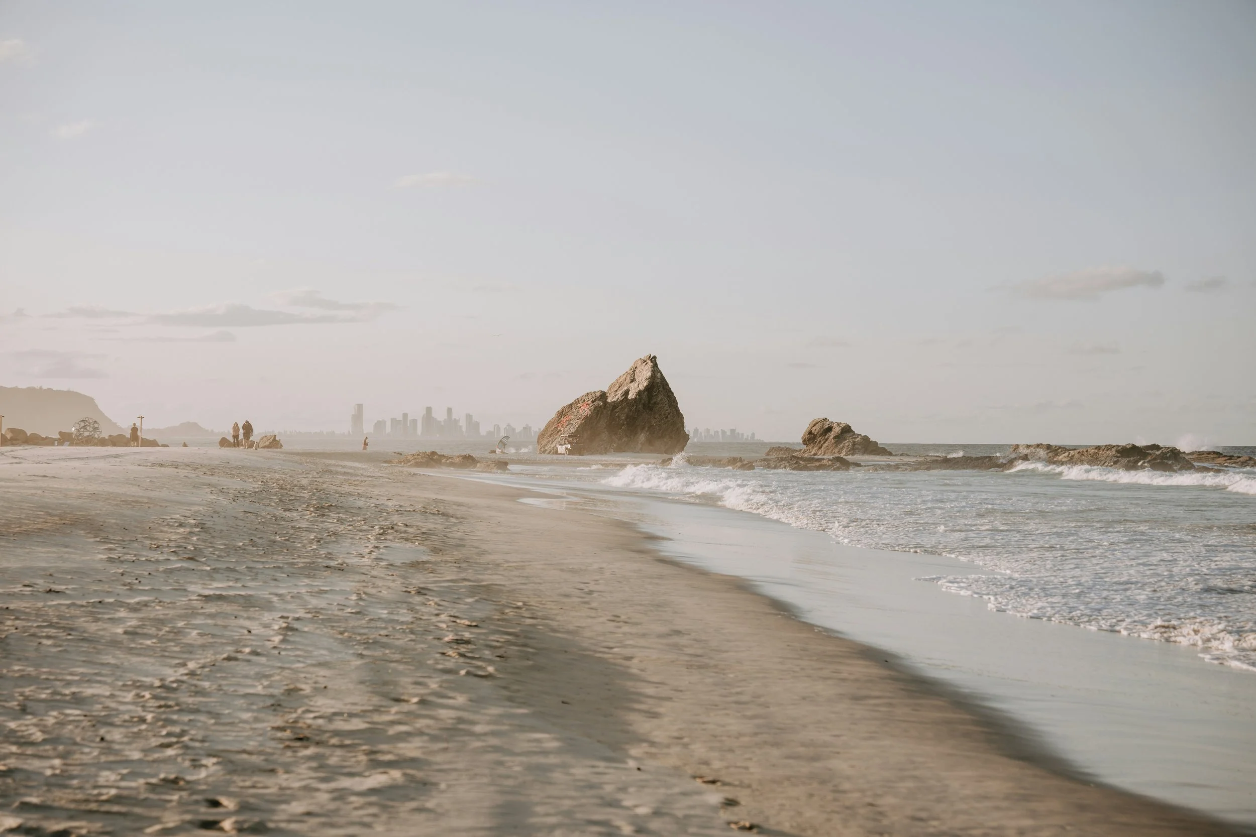 A quiet beach with footprints in the sand, large rocks in the ocean, and a city skyline in the distance under a cloudy sky.