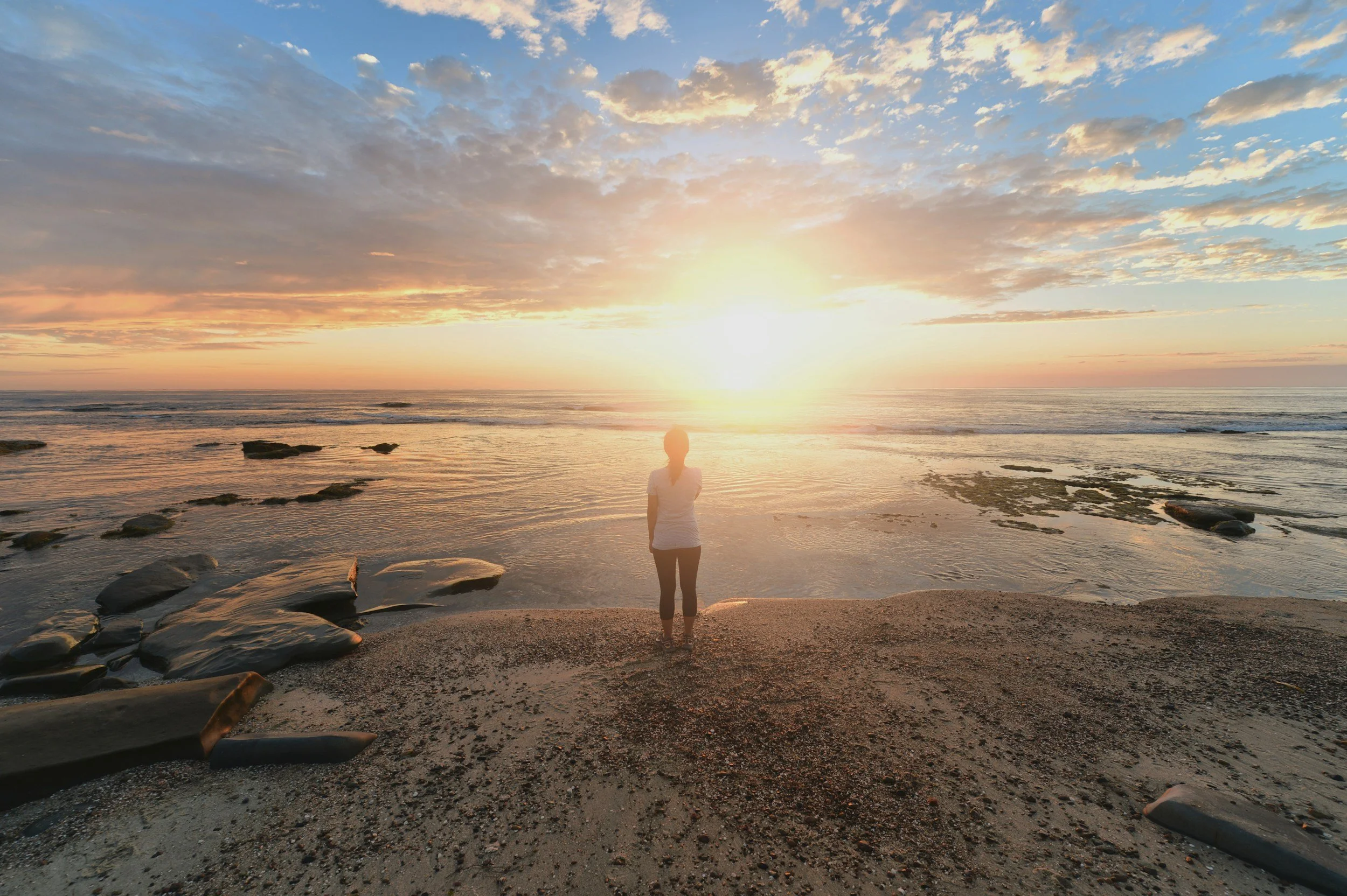 Person standing on the beach at sunrise, facing the ocean with waves and clouds in the sky.