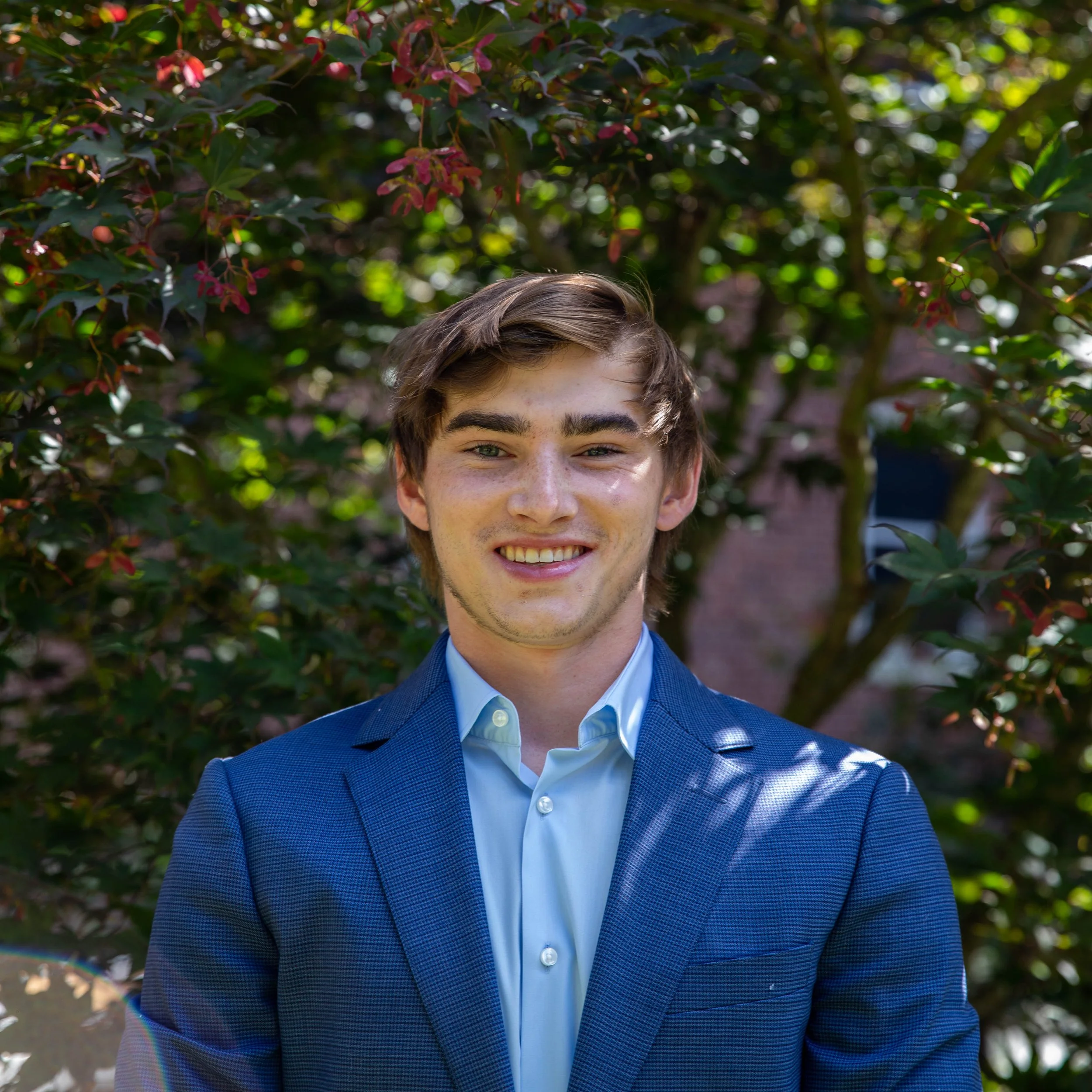 Young man in a blue suit smiling outdoors in front of green foliage.
