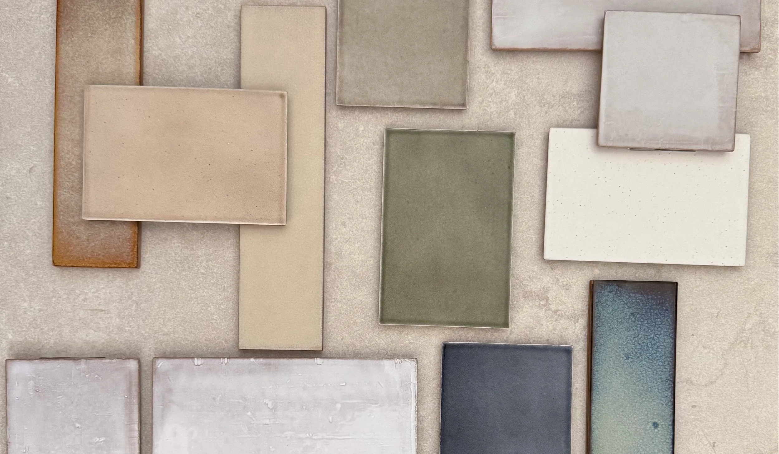 Arrangement of various rectangular ceramic tiles in muted tones including beige, cream, gray, green, white, and a patterned tile with blue and green hues.