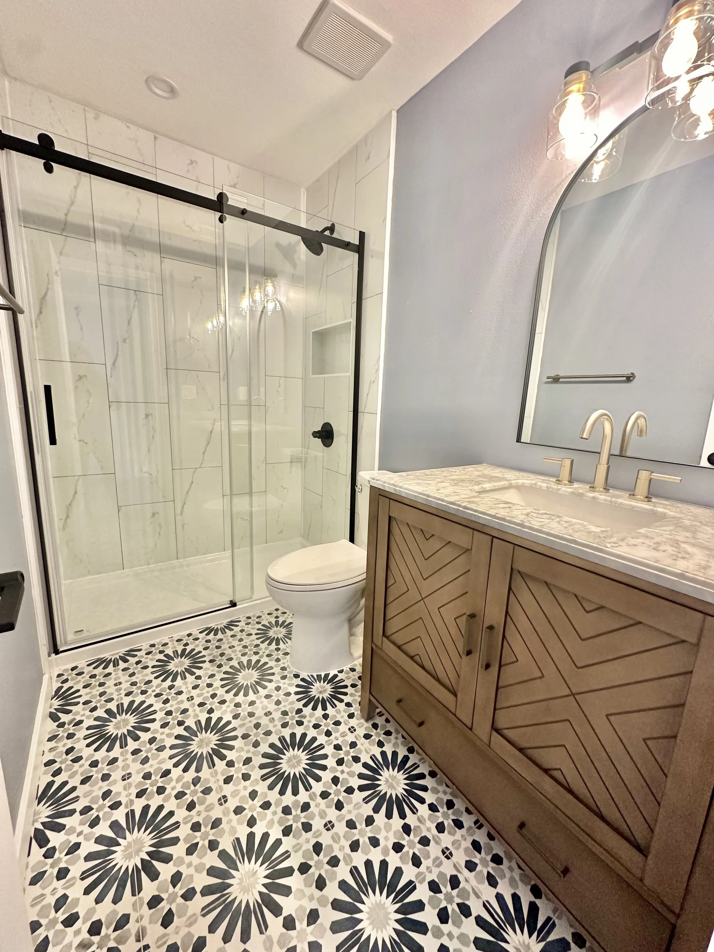 A bathroom with a patterned tile floor, a glass shower enclosure with marble tiling, a wooden vanity with a marble countertop, and a mirror above the sink, illuminated by round light fixtures.European Design. Interior Design. Dallas. Highland Park.