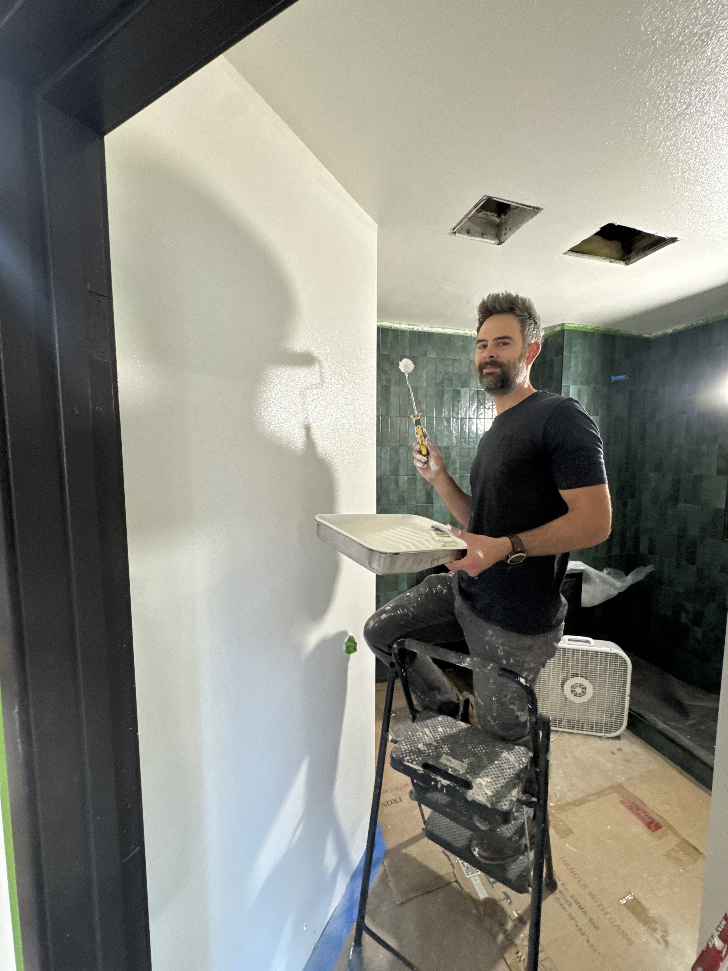 A man with a beard and short hair, wearing a black t-shirt and paint-splattered pants, stands on a step stool painting a wall white. He holds a tray with paint in one hand and a roller brush in the other. The ceiling has two open rectangular air vents and the room has dark green tiled walls and cardboard on the floor.