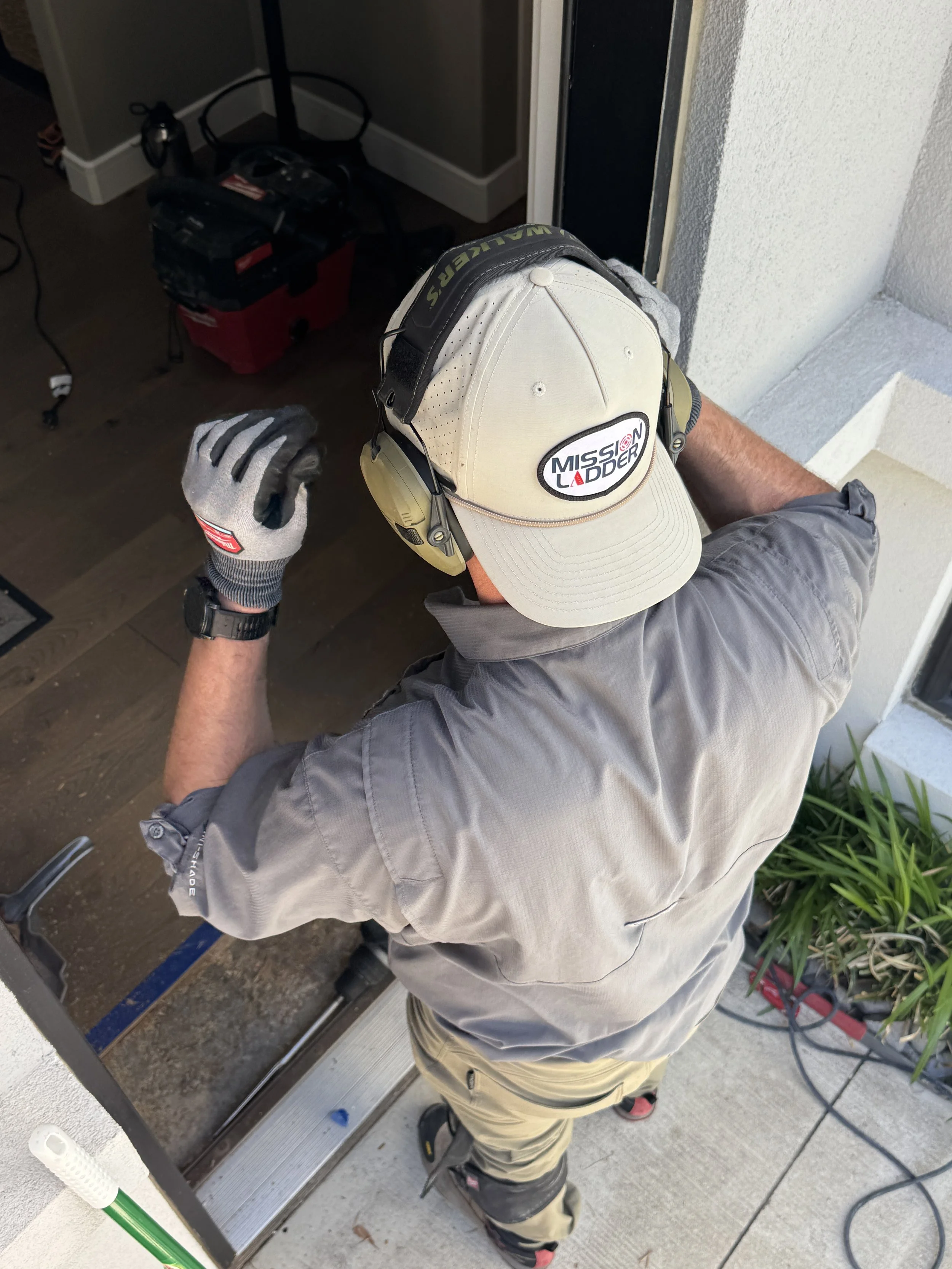 Man installing or repairing a door or door threshold, viewed from above, wearing a cap with 'MISSION LADDER' logo, work gloves, and outdoor work pants.