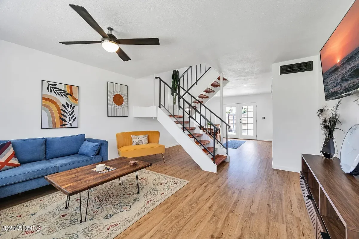 Living room with hardwood floor, blue sofa, yellow accent chair, wooden coffee table, wall art, a ceiling fan, and a staircase with dark railing leading upstairs. There is a TV mounted on the wall and a cabinet with decorative items.