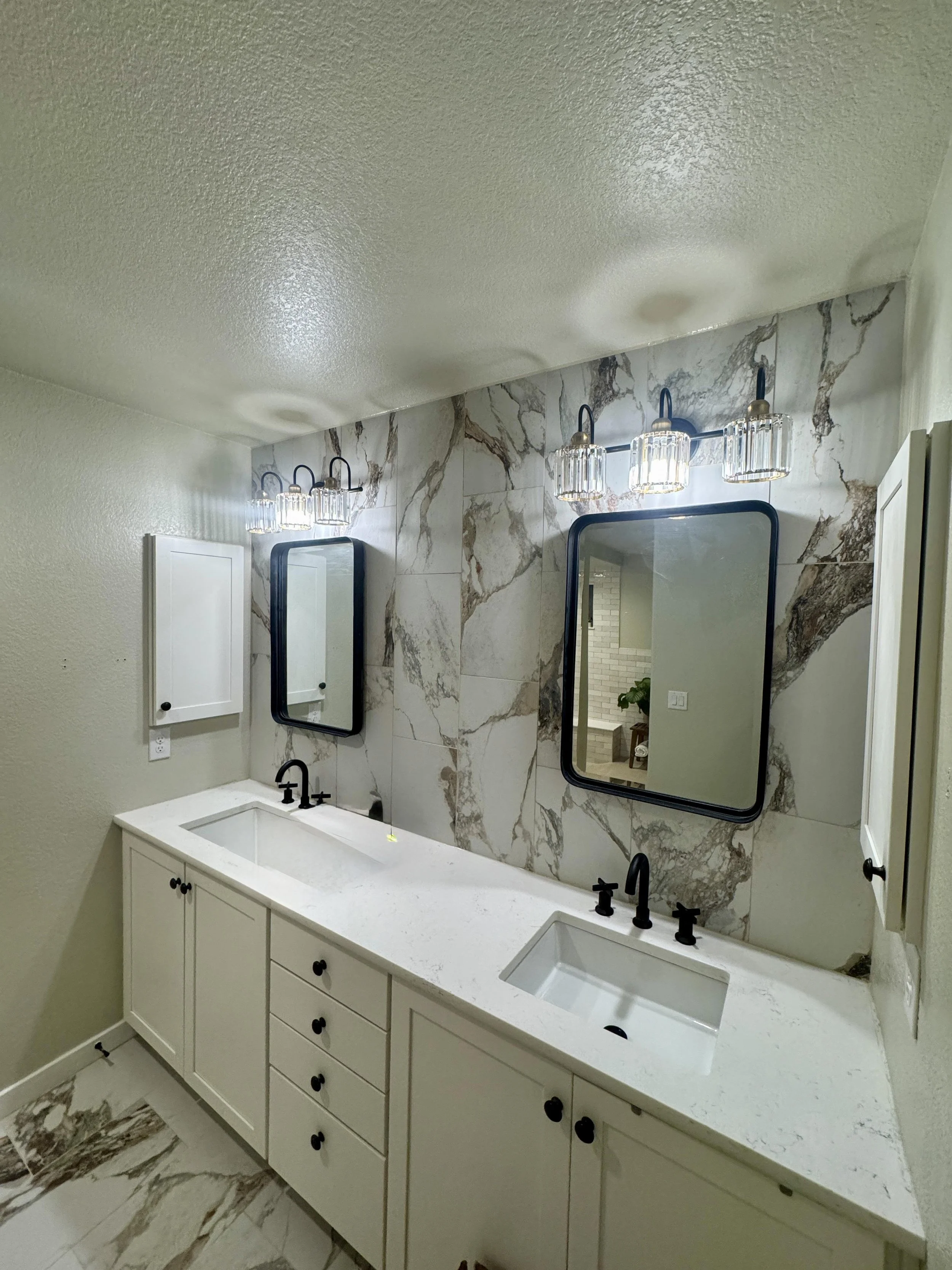Modern bathroom vanity with dual sinks, black fixtures, white cabinetry, large marble wall behind, and two rectangular mirrors with black frames. Two wall-mounted light fixtures with glass shades are above each mirror.European Design. Interior Design