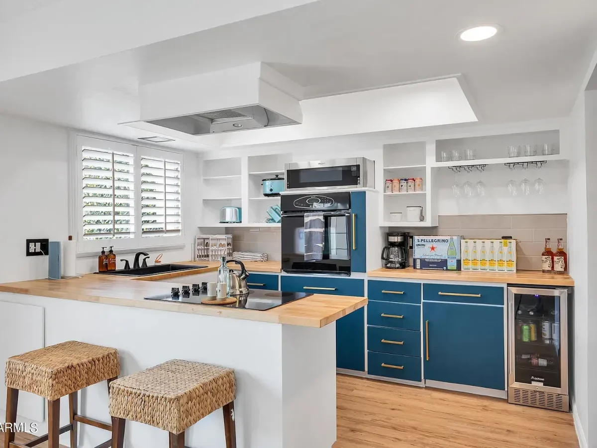 Modern kitchen with white walls, wooden countertops, blue lower cabinets with gold handles, and open shelving with glasses, cups, and spices. There is a microwave, coffee maker, and small wine fridge. Two woven stools are at the counter with a window