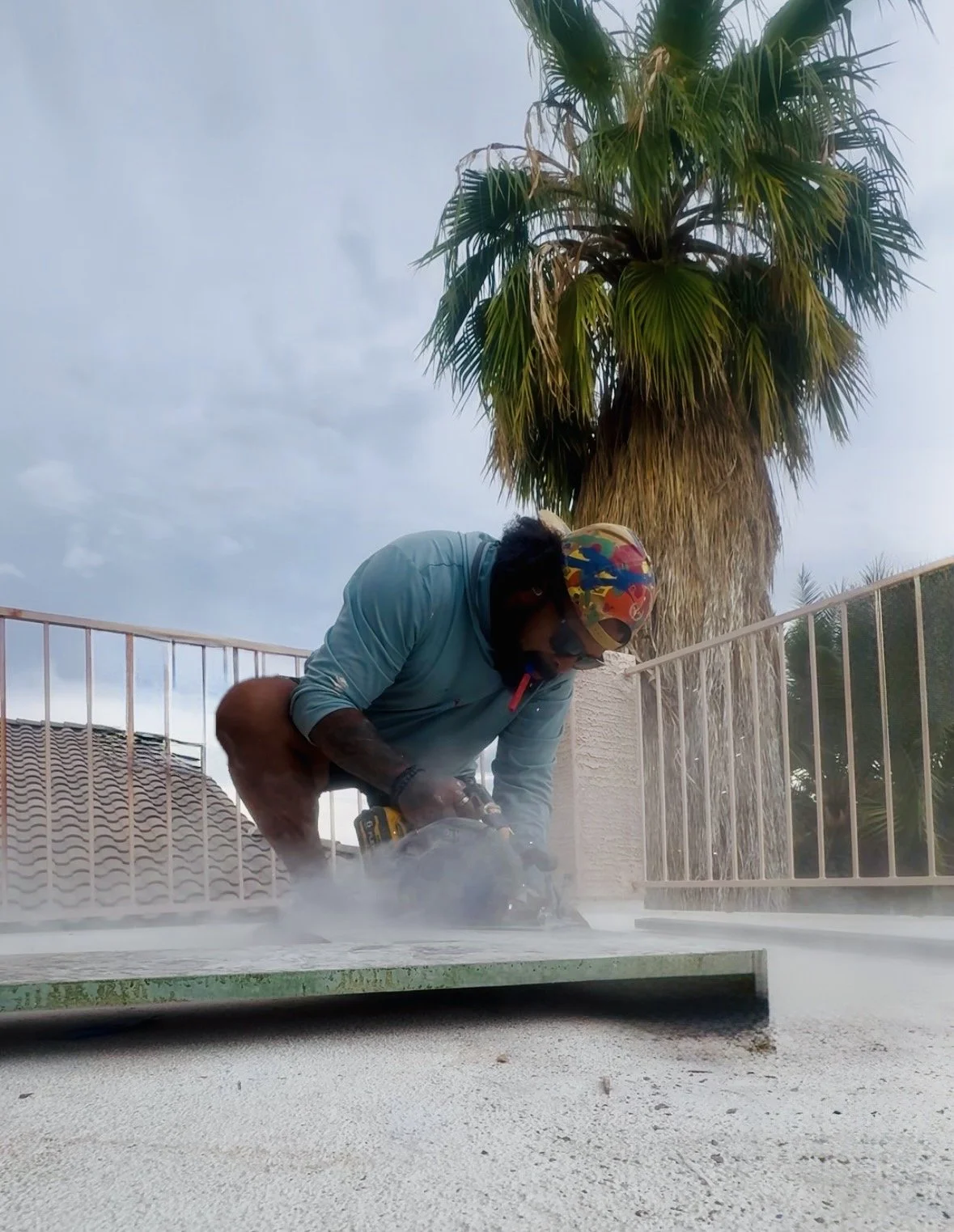 Man using a power drill on a white surface outdoors, with a palm tree and cloudy sky in the background.