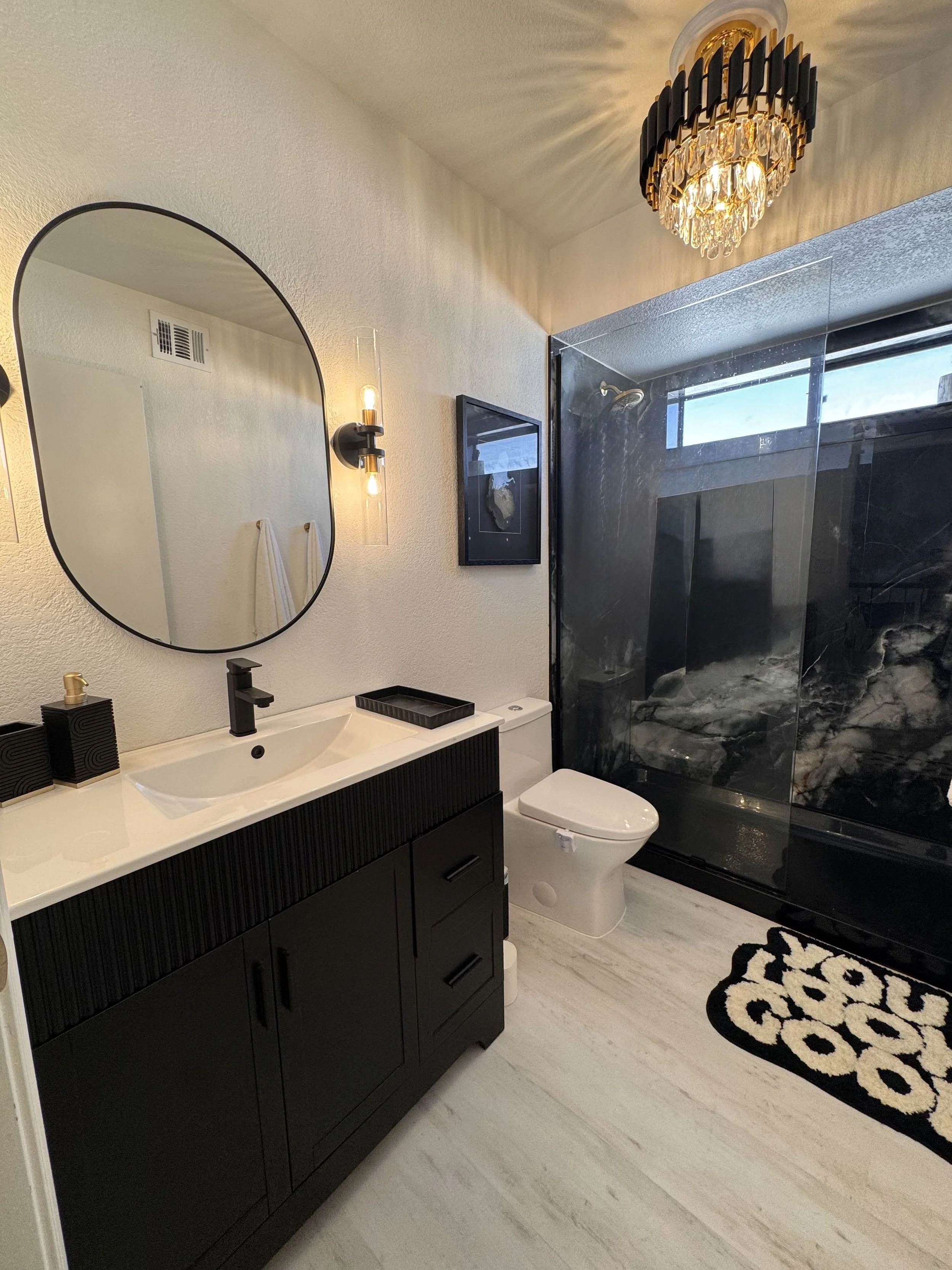 Modern bathroom with black vanity, oval mirror, black sink faucet, wall-mounted light fixture, framed wall art, toilet, black and white patterned rug, and a black marble shower with glass door and a window.