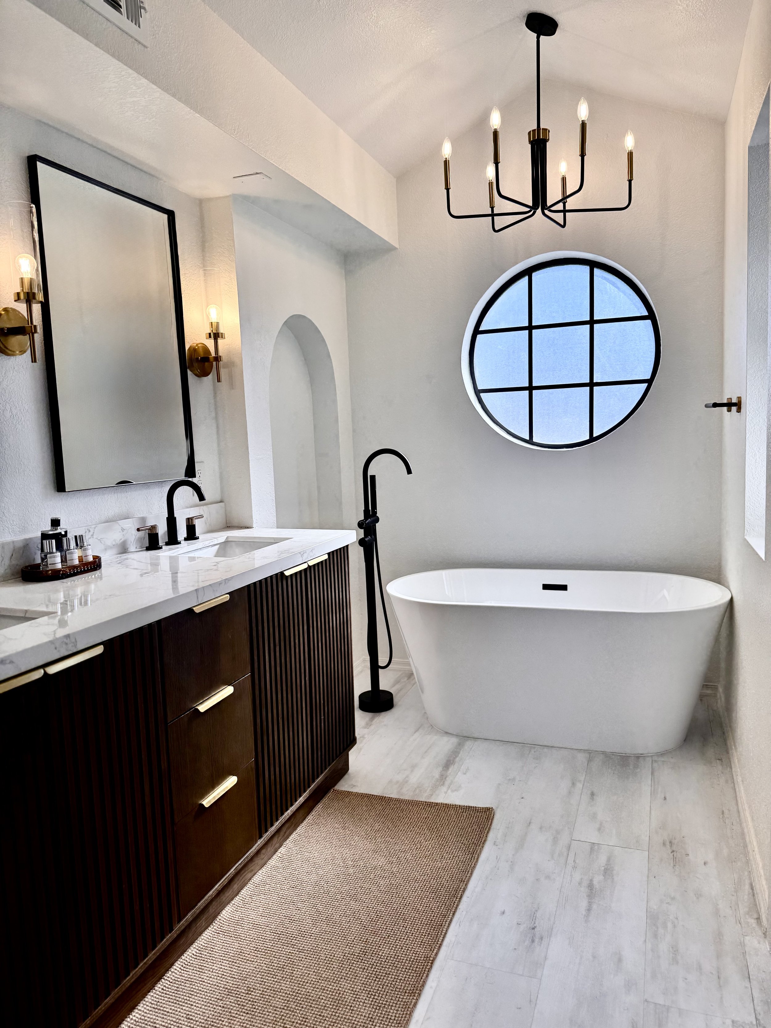 Modern bathroom with white bathtub, arched window, double sink vanity with dark wood cabinets, large mirror, and black fixtures, illuminated by a chandelier and wall sconces.European Design. Interior Design. Dallas. Highland Park. North Dallas. 