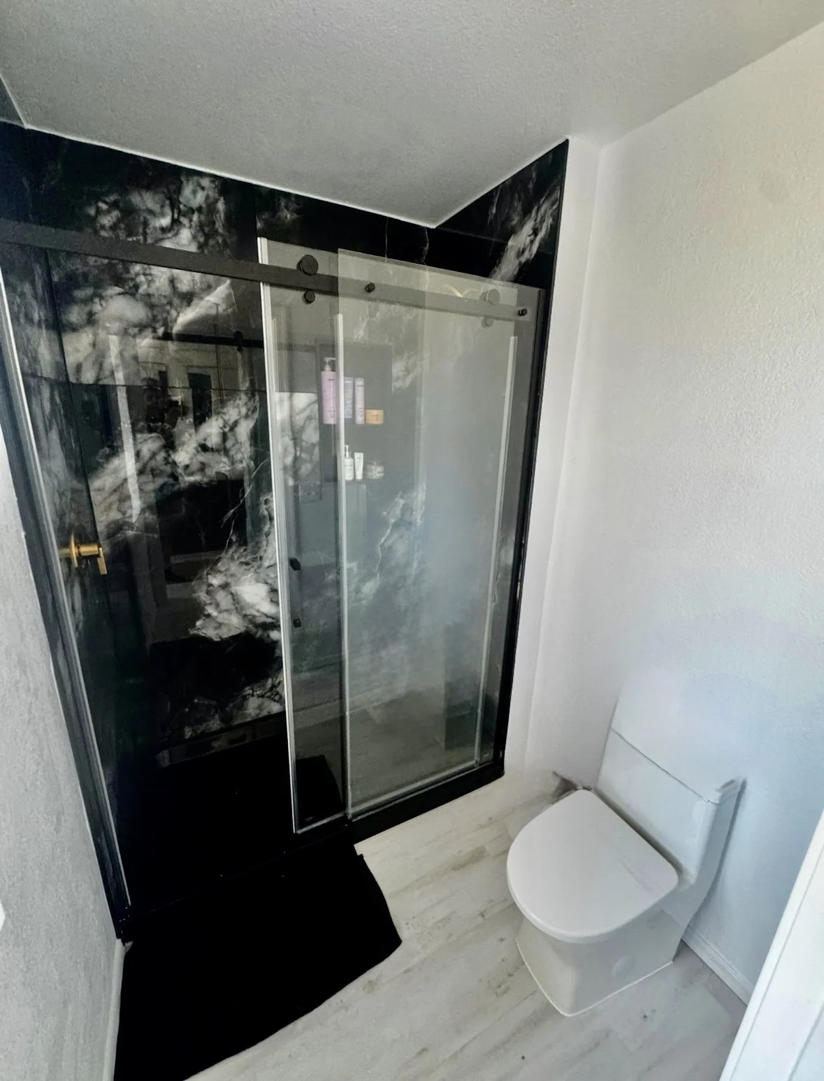 Black-and-white marble shower enclosure with glass sliding doors, a white toilet, and oak flooring. XL porcelain slab. 