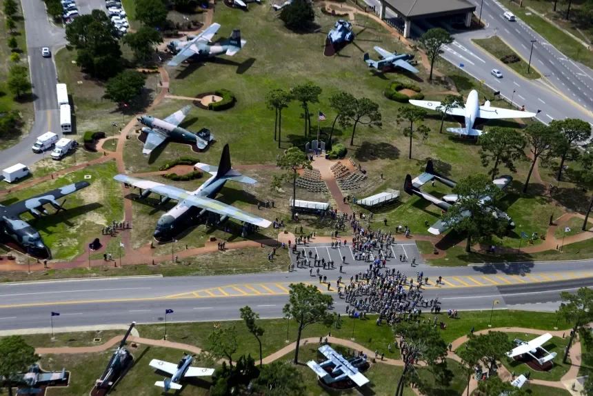 An aerial view of a park with multiple aircraft exhibits and a crowd of people gathered near the stairs leading to the park entrance. The park is surrounded by roads and greenery.