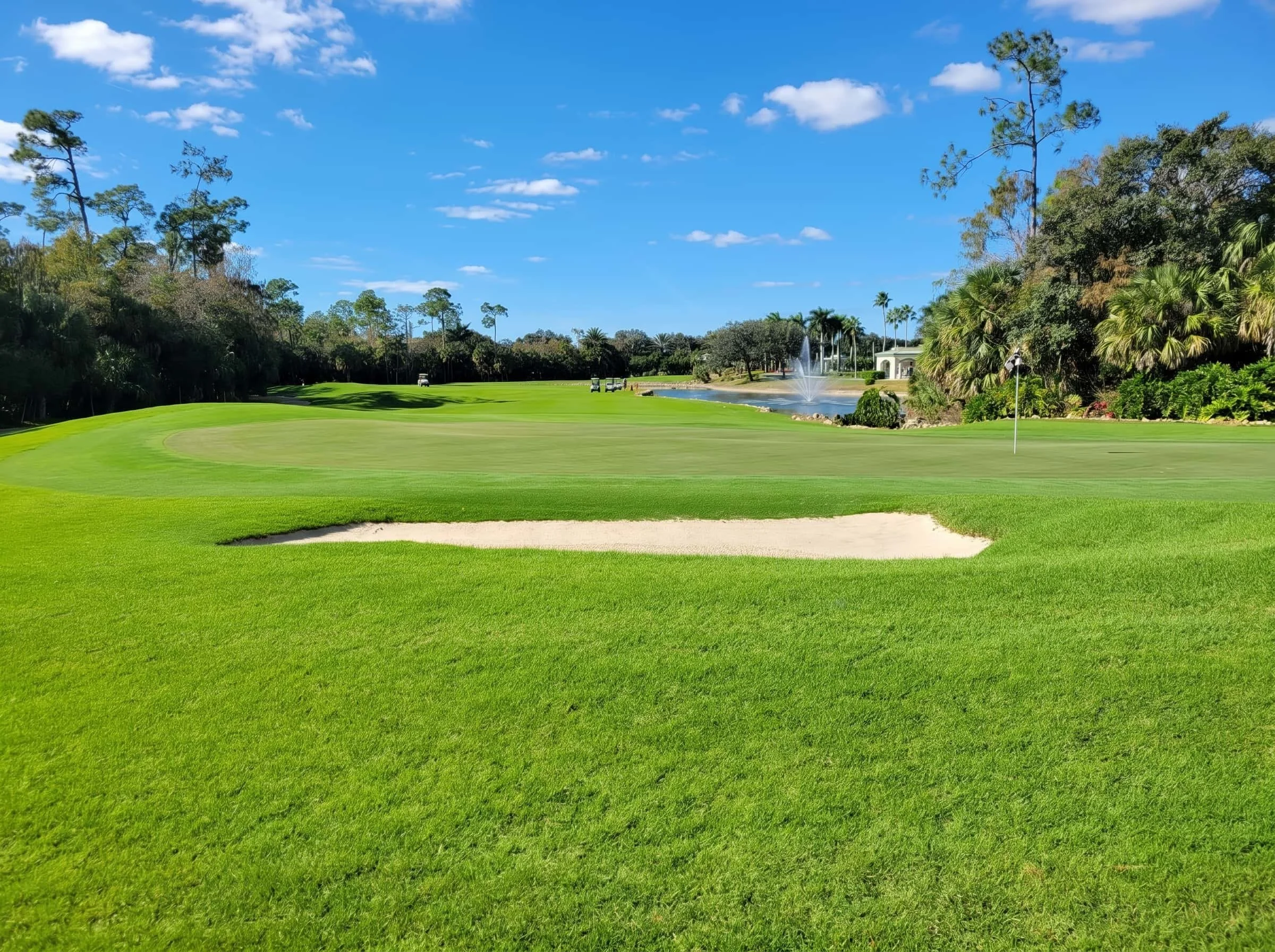 A lush green golf course with a sand trap, a water fountain, and a flag on the putting green under a bright blue sky with scattered clouds.