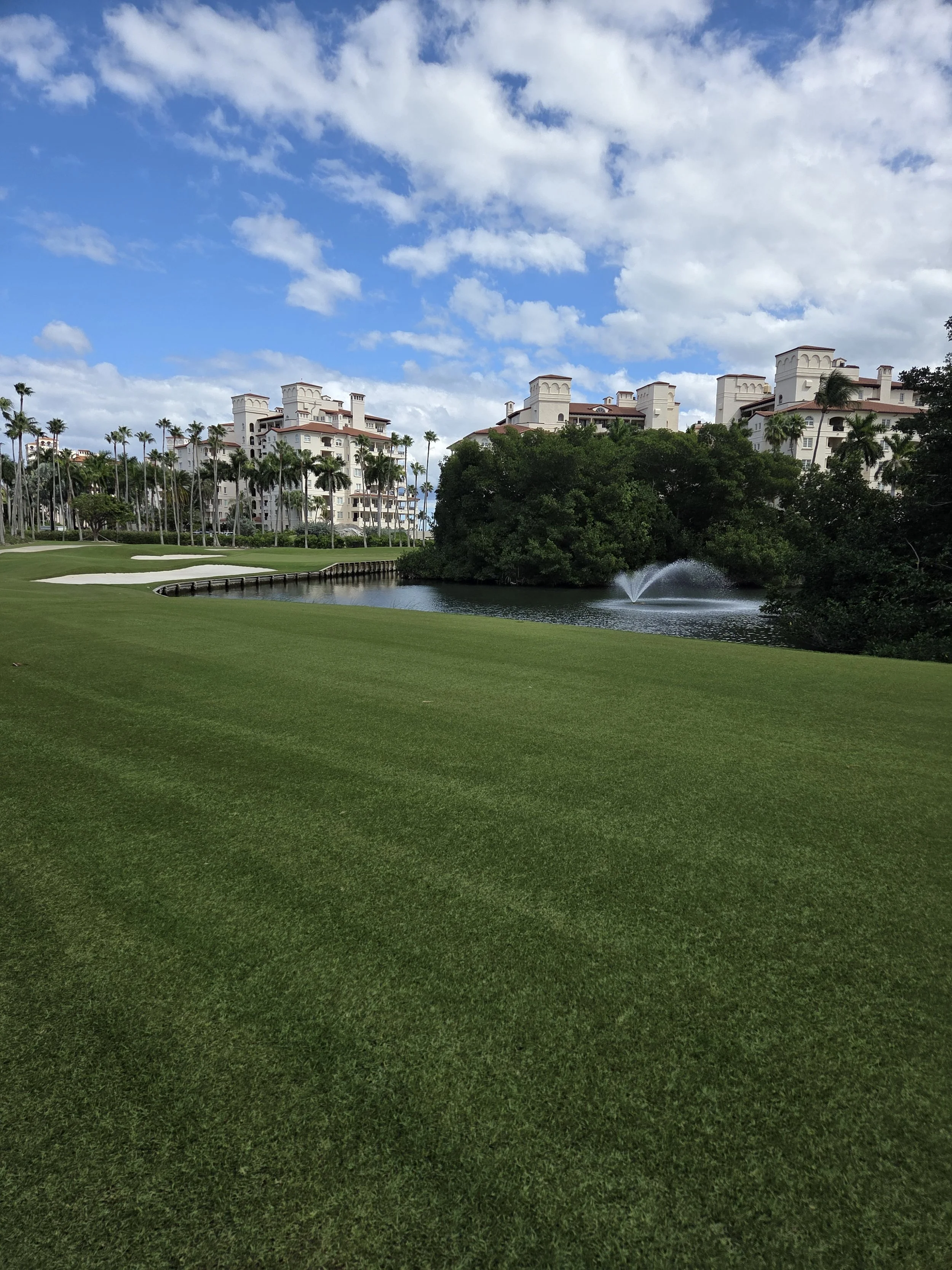 A golf course with well-maintained green grass, a water hazard with a fountain, surrounded by lush trees and palm trees, with white residential buildings in the background under a partly cloudy sky.