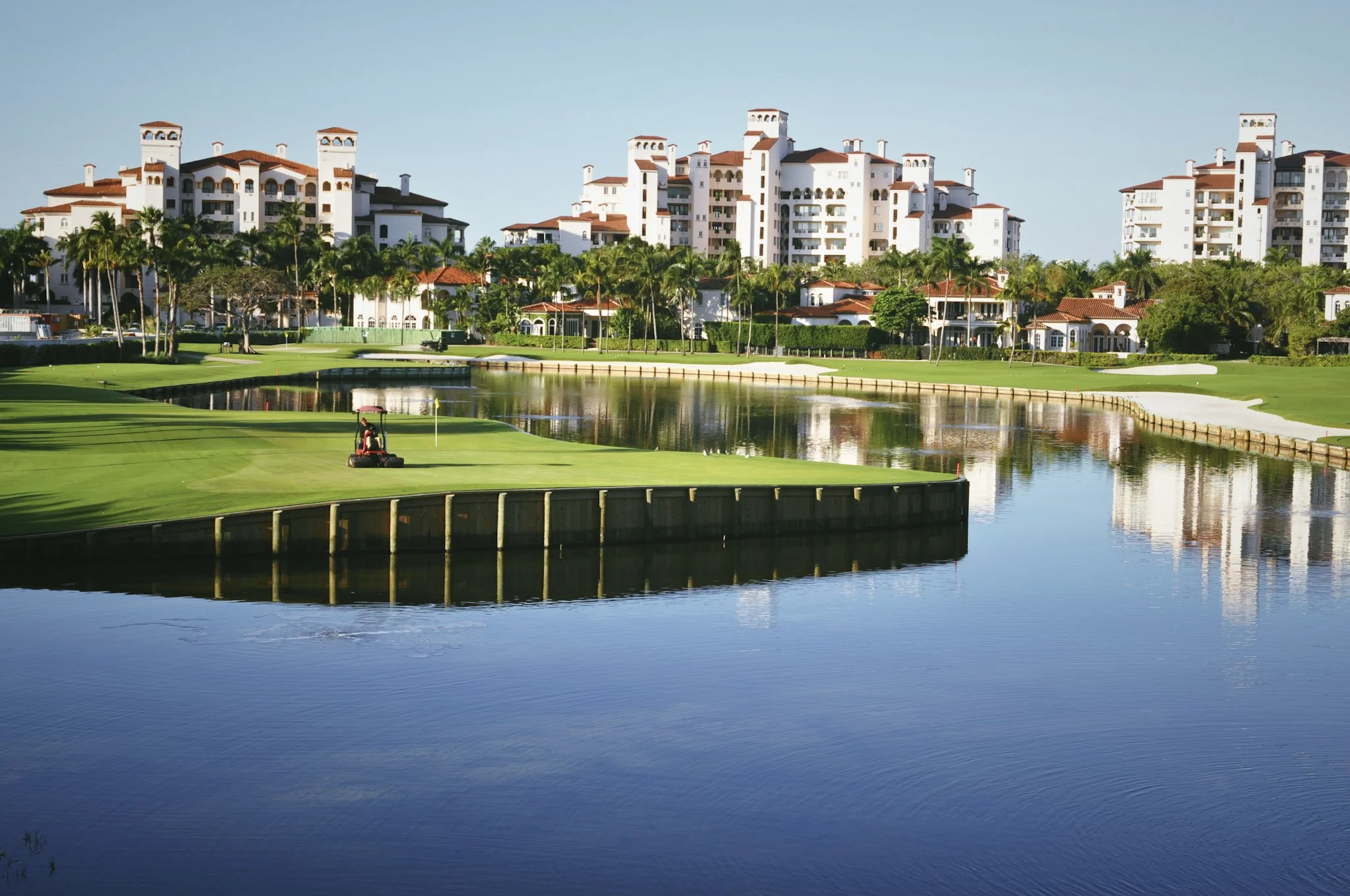 A golf course with a putting green and a person riding a golf cart near a pond, with a large white residential building with red roofs in the background under a clear blue sky.