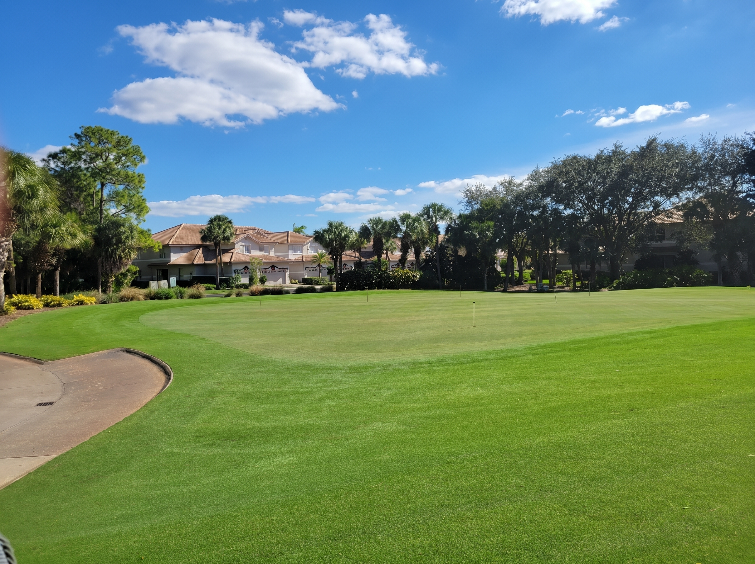 A golf course with manicured green grass and several small flags, surrounded by trees and residential houses in the background, under a blue sky with scattered white clouds.
