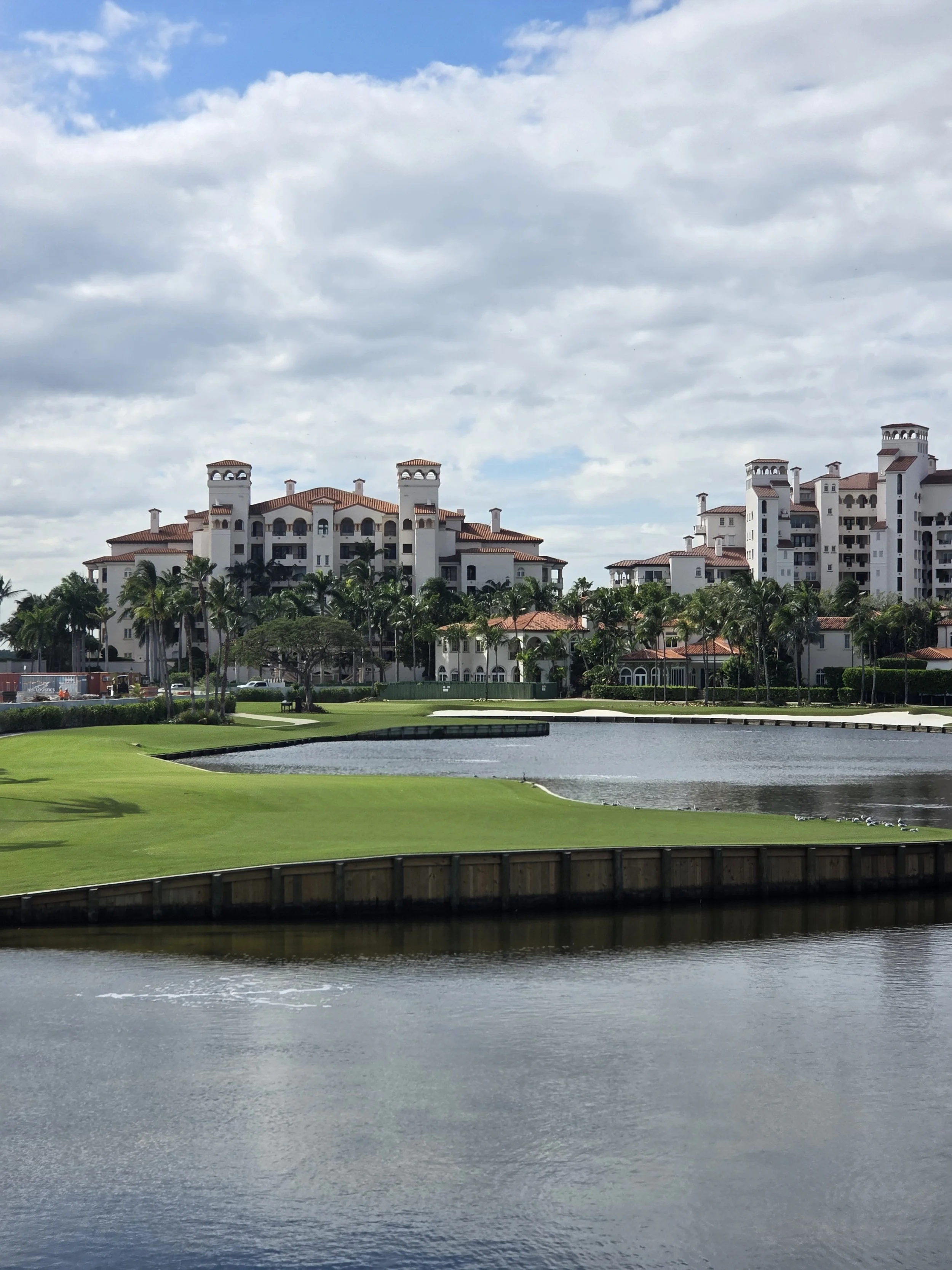 A large white residential building with red tile roofs and multiple towers sit behind a row of palm trees, with a pond or small lake and well-manicured green lawn in the foreground. The sky is partly cloudy with patches of blue.