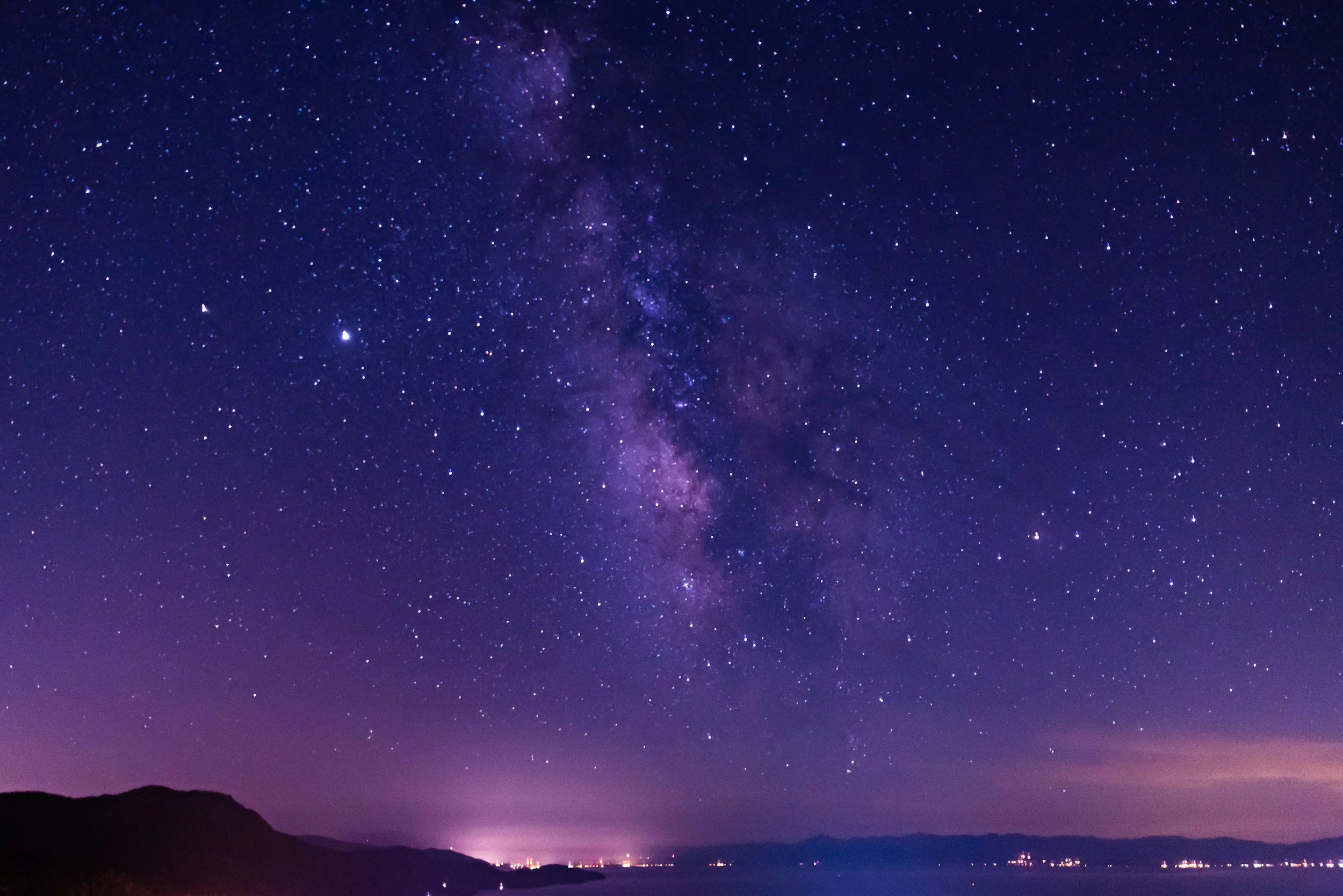 Clear night sky filled with stars and the Milky Way galaxy over a dark silhouette of hills and distant city lights at the horizon.