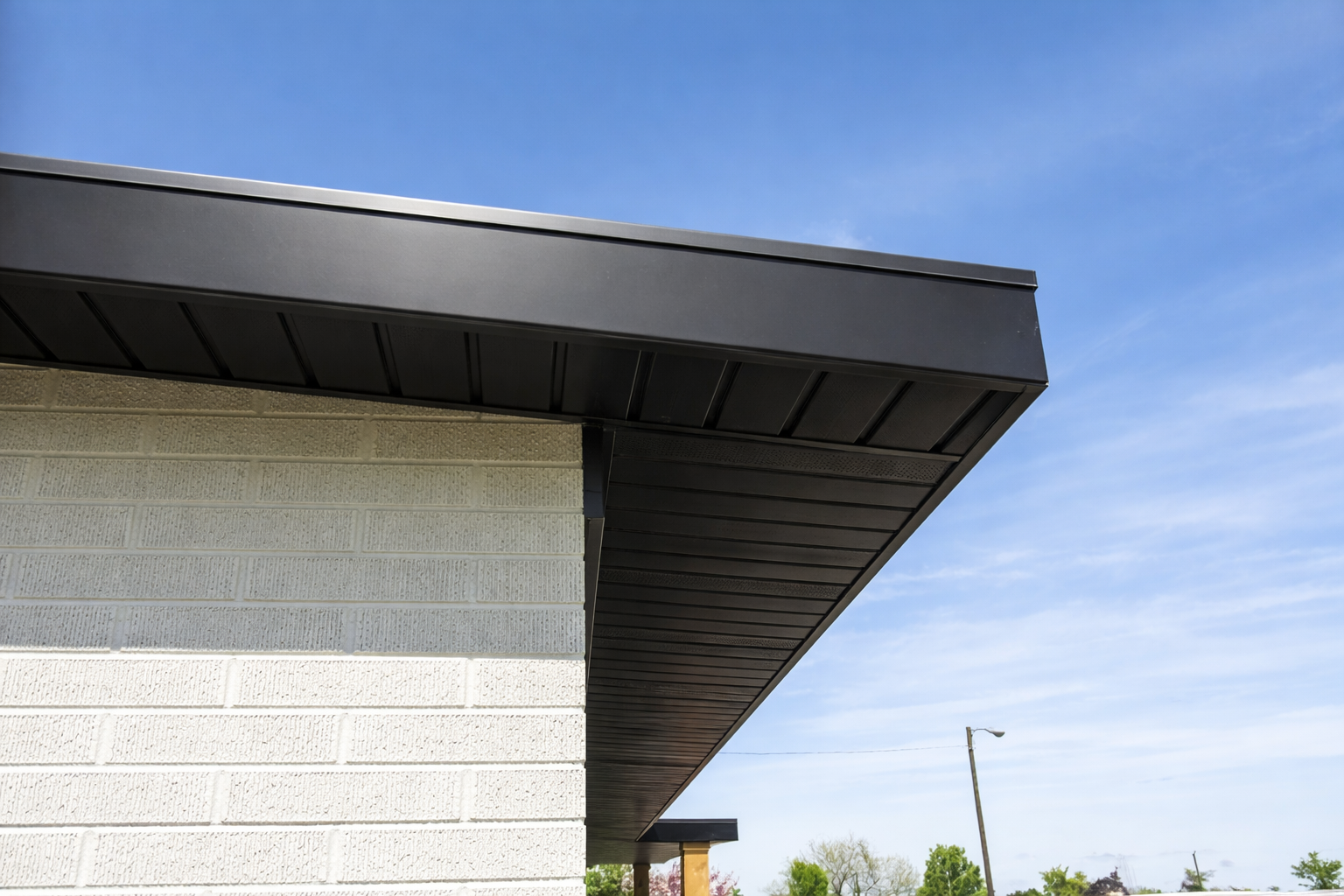 Close-up of a building's corner showing a brick wall and an overhanging black metal roof, with a background of a blue sky and a streetlight.