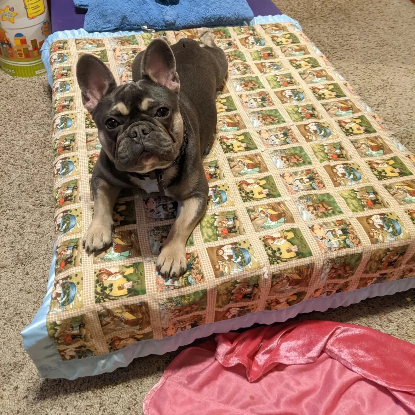 A French Bulldog puppy lying on a colorful, patterned bedspread with various scenes. There are cushions and a soft blue blanket behind the puppy, and a pink, silky cloth at the edge of the bed.