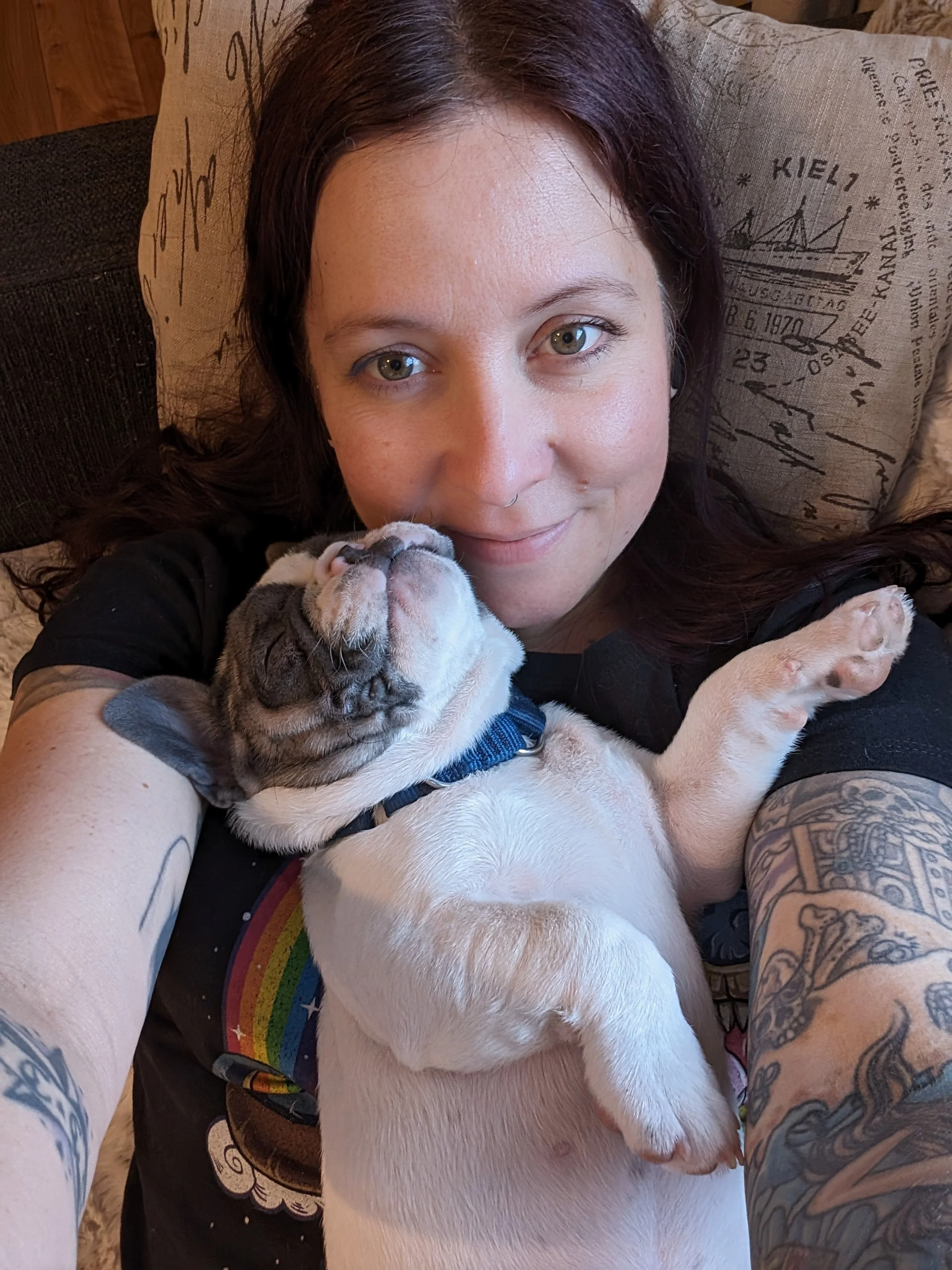 A woman with dark hair and tattoos on her arms holding a sleeping French Bulldog puppy with a rainbow on her shirt. They are lying on a couch with a pillow in the background.