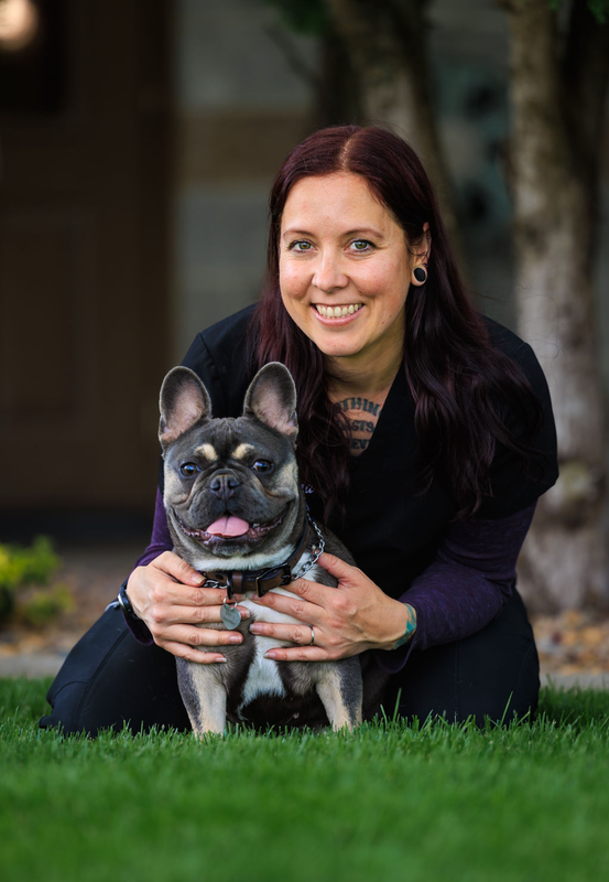 A woman with long dark hair and tattoos kneeling on grass, smiling, holding a small French Bulldog with a happy expression and tongue out.