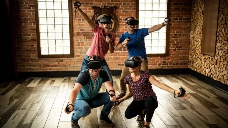 Five people wearing virtual reality headsets and holding controllers in a brick-walled room with windows.