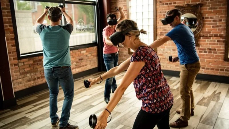 Four people in a room with brick walls and wooden floors, each wearing virtual reality headsets and holding controllers, engaging in a VR activity.