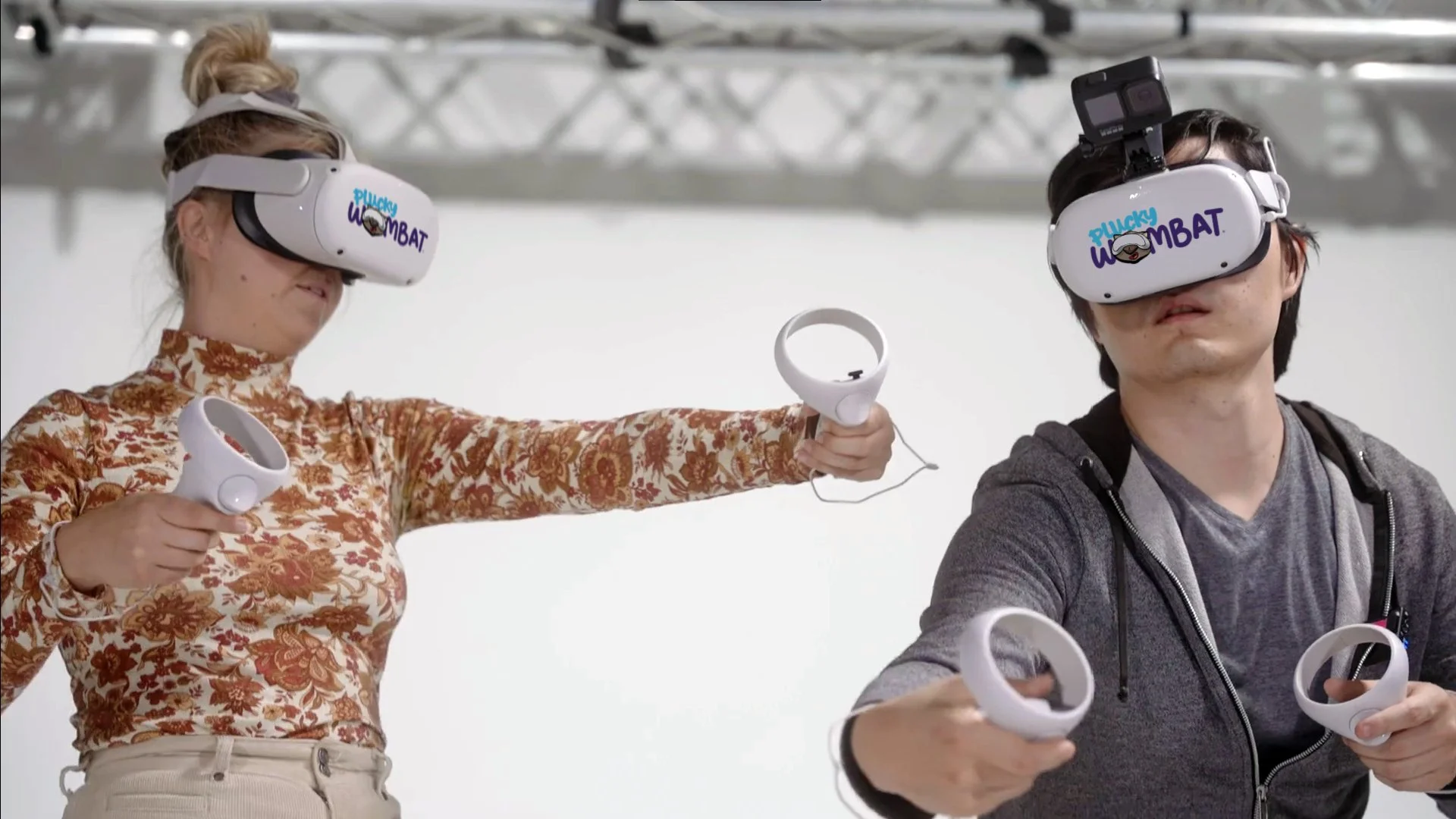 Two people wearing virtual reality headsets and holding VR controllers, engaging in a virtual reality experience. The woman on the left has a floral shirt and beige pants, and the man on the right has a gray hoodie. They are indoors with a neutral background.