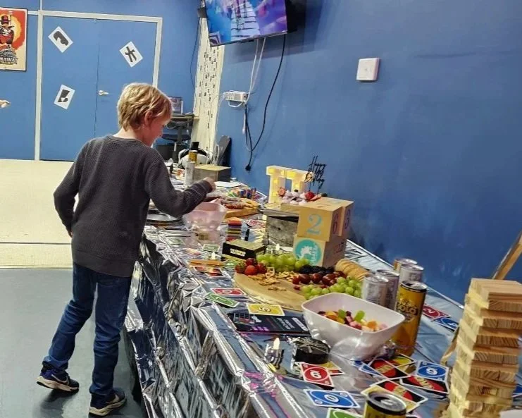 A young person standing in front of a table filled with snacks, drinks, and games in a blue-walled room with decorations. a birthday party at DropBear HQ Pakenham
