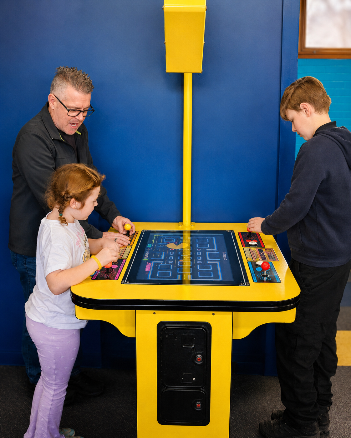 Two children, a girl and a boy, playing Pac Man Battle Royale arcade game with a man also playing, in an indoor game room with blue walls. at DropBear HQ Arcade in Pakenham