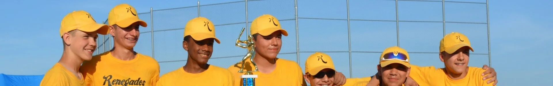 A group of young baseball players in yellow uniforms and caps celebrating on a baseball field, holding a trophy, with a blue sky and chain-link fence in the background.