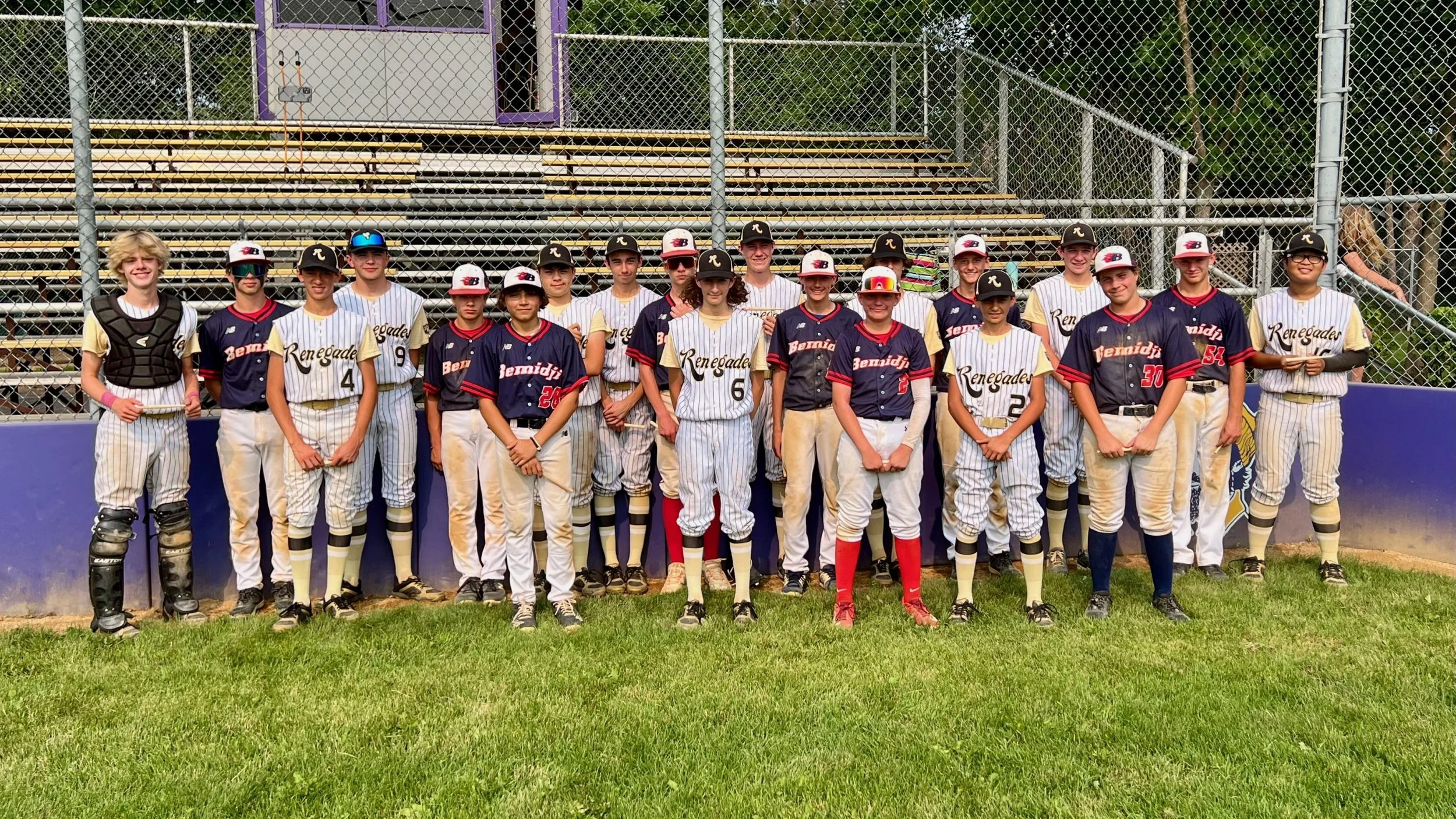 A group of young baseball players and their coach posing for a team photo on the field, with a chain-link fence and empty bleachers in the background.