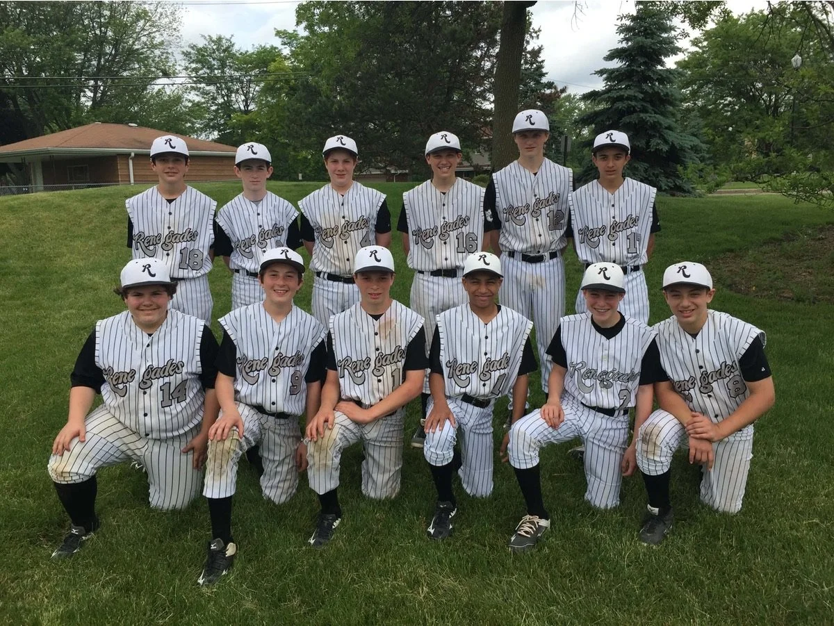 Youth baseball team in black and white striped uniforms posing outdoors on a grassy field with trees and houses in the background.