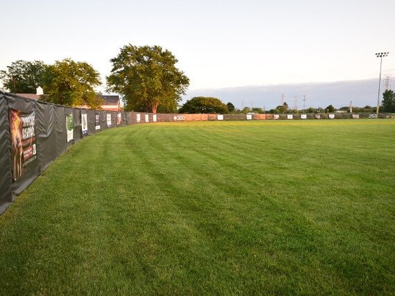 Empty baseball or softball field with green grass, outfield fence with sponsor banners, trees, and stadium lights in the background.