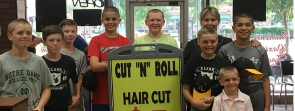 Group of children and a woman standing behind a yellow and black sign that reads 'Cut 'N' Roll Hair Cut' inside a store or salon.